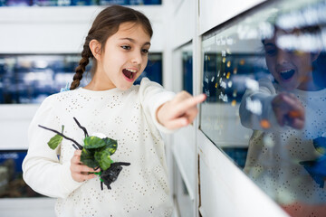Portrait of interested cute girl pointing with finger on aquarium with tropical fishes in pet shop