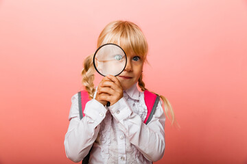 Portrait of a cute little girl holding a magnifying glass near her eye on a pink background.