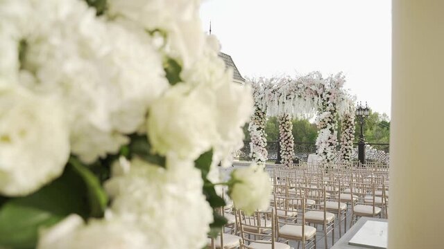 Luxurious square wedding arch with branches of blossoming cherries and roses on terrace. Mirror path leads to arch on both sides of which there are rows of chairs. In foreground is flower arrangement