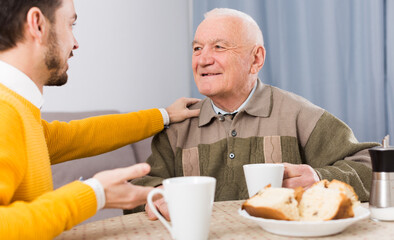 Smiling elderly father and son having breakfast and warmly talk at table at home