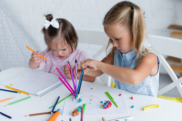 blonde girl reaching color pencils near disabled toddler kid with down syndrome in private kindergarten