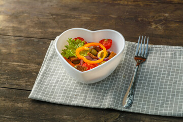 Vegetable salad in a heart shape plate on a wooden table on a checkered napkin next to a fork.