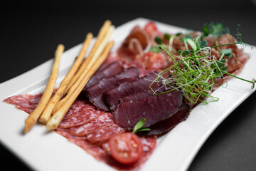cold meat plate with salami, prosciutto and bread sticks on black background