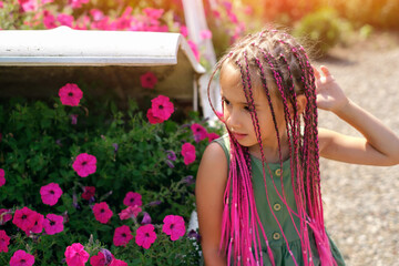 A beautiful cute girl with pink Afro-pigtails from kanekalon stands in front of a flower bed with...