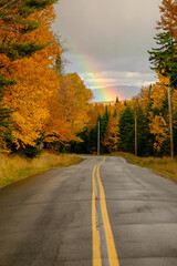 A lonely empty highway through colorful autumn trees and a rainbow at the end of the road
