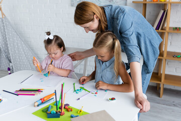 teacher looking at preschooler girl and disabled child with down syndrome molding plasticine in...
