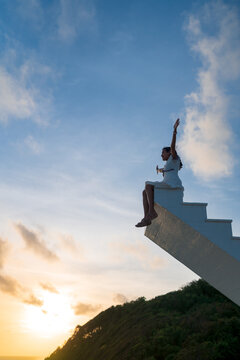 Asian woman raise arms up sit on top of wooden staircase with sunset sky in the background, victory, success concept 
