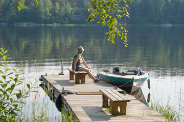 Landscape with a lake shore. There is a wooden pier with a moored boat. A woman sits on a bench and looks into the distance.