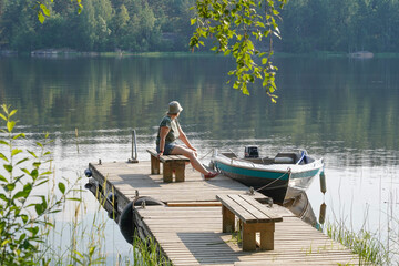 Landscape with a lake shore. There is a wooden pier with a moored boat. A woman sits on a bench and...