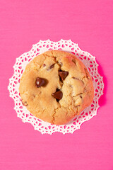 Close up of a homemade chocolate chip cookie on a bright pink background with a white doily 
