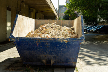 Blue dumpster with broken bricks and plaster after apartment repair on a sunny summer day. Large container with construction debris on the street. Removal of construction waste.