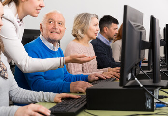 Enthusiastic senior man taking computer lessons from young female instructor with group of elderly people..