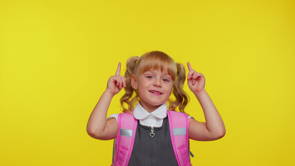 Attractive blond teenage girl kid in school uniform wears pink backpack showing thumbs up and pointing up at copy space for promotional content on yellow studio background. Education, back to school