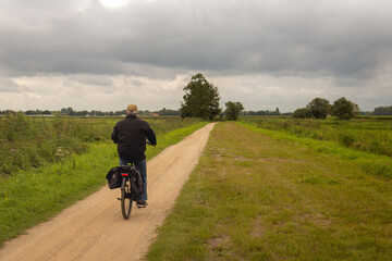 Old man in a cap is cycling on a straight sandy path in a nature reserve in a Dutch polder in the...