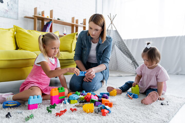 kindergarten teacher playing building blocks with preschooler girl and toddler kid with down syndrome