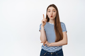 Excited young woman got an idea, raising finger eureka gesture, pointing up, find or discover something interesting, standing in striped tshirt over white background