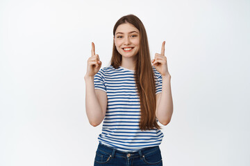 Young happy woman showing information, pointing fingers up and smiling pleased, demonstrating sale banner, standing over white background