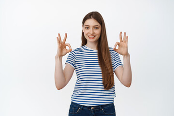 Smiling girl shows okay signs in approval, stands over white background in casual clothes