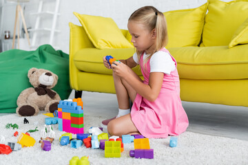blonde girl in pink dress playing building blocks on carpet in kindergarten playroom