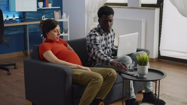 Interracial couple of parents expecting baby sitting on sofa at home. Young black husband using laptop while helping pregnant caucasian wife with TV remote control in living room.