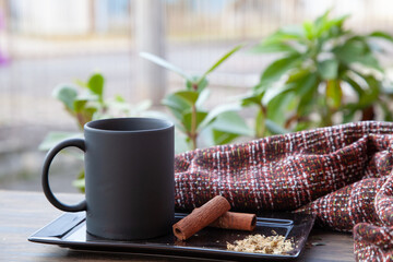 lack mug with tea, cinnamon and plaid blanket on a small table on the balcony with potted plants in the blurred background