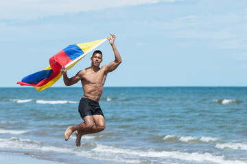 Man holding a Venezuelan flag while jumping on the seashore at the beach.