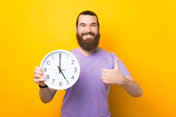 Happy bearded man is holding a round clock and showing thumb up.