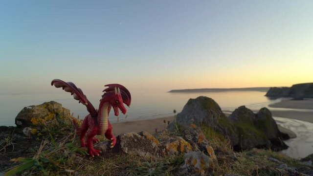 Red toy dragon overlooking Three Cliffs Bay at sunset, the Gower peninsula, Swansea, South Wales, United Kingdom. Symbol of Wales and Welsh landscape. Popular destination with beautiful beach.