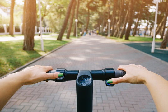 Young woman drives an electric scooter in a city park. Point of view on the steering wheel with female hands.