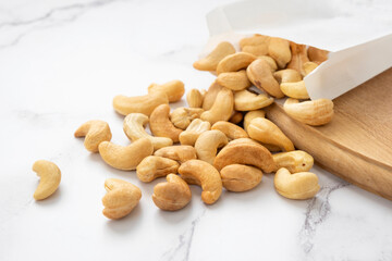 Many cashew nuts on wooden board on marble background out of white paper packet. Source of natural fats and protein. Healthy eating or diet concept.