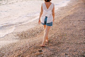 Barefoot woman walks along the pebble beach near the water. Back view