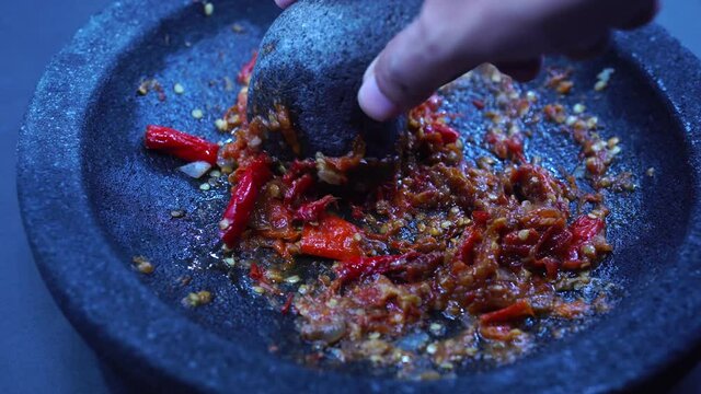 The hand is grinding the red chili sauce in a stone mortar. Chef preparing Indonesian traditional spicy hot chili sauce