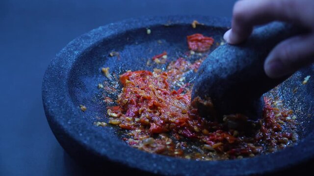 The hand is grinding the red chili sauce in a stone mortar. Chef preparing Indonesian traditional spicy hot chili sauce. Zoom out