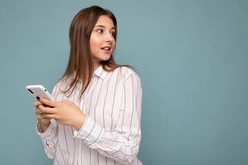 Photo of beautiful young blonde woman wearing casual white shirt isolated over blue background wall holding smartphone and texting message looking to the side