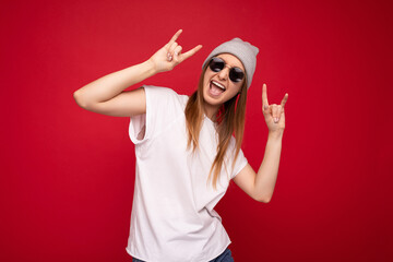 Fototapeta na wymiar Photo shot of beautiful positive young dark blonde woman wearing casual clothes and stylish sunglasses isolated over colorful background wall looking at camera having fun and showing rock and roll