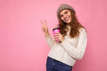 Attractive young positive blonde woman wearing casual stylish clothes isolated over colorful background wall holding paper cup for mockup drinking coffee looking at camera and showing peace gesture