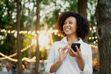 A beautiful Afro-looking woman types a message on her phone in a summer cafe. The student does the task online for the new course of the lesson.