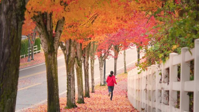 Teenage girl jogging along sidewalk in beautiful autumn foliage while leaves falling from the tree on a windy day