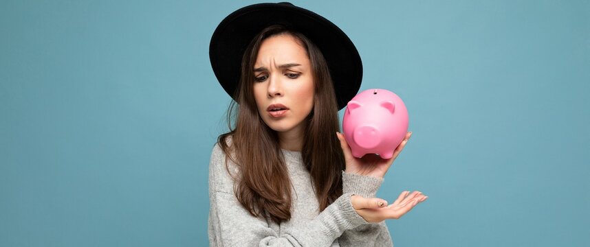 Panoramic portrait of dissatsfied sad upset young beautiful brunette woman with sincere emotions wearing stylish clothes and black hat isolated over blue background with empty space and holding pink