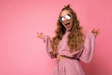 Photo shot of beautiful amazed shocked young dark blonde woman wearing casual clothes and stylish sunglasses isolated over colorful background looking at camera with open mouth