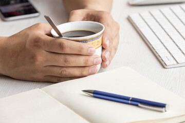 Fototapeta na wymiar workplace with a person's hands holding a cup with hot coffee, next to it an open notebook with blank sheets and a pen, in the background a desk