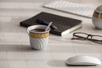 wooden desk with a cup of coffee with a spoon, it is next to glasses, computer mouse, sugar bowl, notebook, pen and keyboard, detail of objects