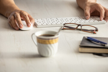 studio with a person working at his desk, detail of his hands next to a modern keyboard, glasses, pen, cup of coffee, computer mouse, keyboard and notebook, lifestyle with office objects
