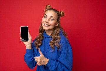 Naklejka premium Attractive smiling young blonde woman wearing stylish blue hoodie isolated on red background with copy space holding smartphone showing phone in hand with empty screen display looking at camera
