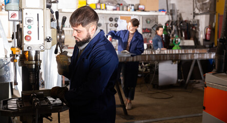 Confident bearded workman drilling metal parts on stationary machine in metalworking workshop ..