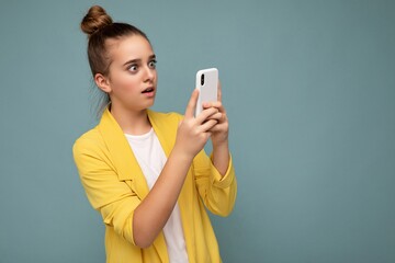 Beautiful amazed young girl wearing yellow jacket and white t-shirt standing isolated over blue background surfing on the internet via phone looking at mobile screen