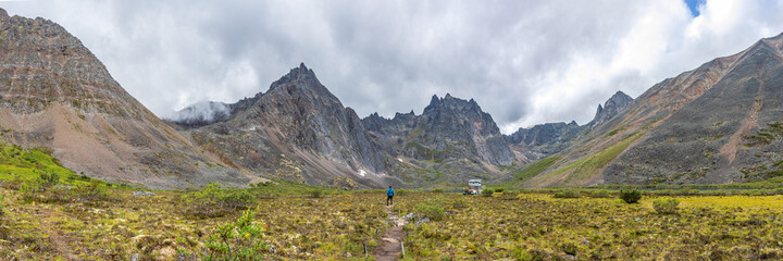 Fototapeta na wymiar Panoramic view of hiker in backcountry campground of Grizzly Lake, Yukon. Amazing towering mountain peaks behind man wearing blue jacket in summer time. 