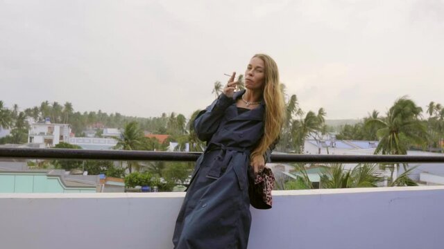 A blonde caucasian girl wearing a dark blue coat is standing on the rooftop in south-east Asia smoking a cigarette