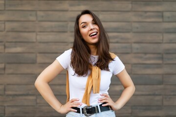Fototapeta na wymiar Portrait of young beautiful positive happy brunette woman standing against brown wall in the street and wearing stylish outfit