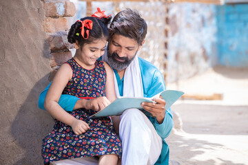 Happy rural indian father helping his daughter in studies, adorable little village girl child holding notebook studying outside home. Man teaching and support female in education.